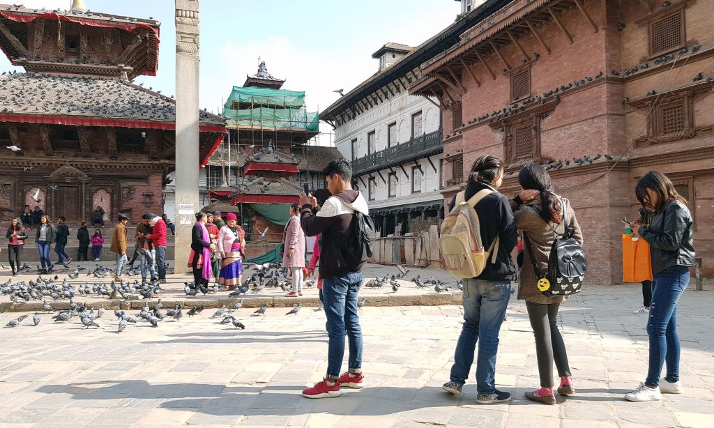 Historiallinen aukio Patanissa, Nepalissa, etualalla seisoo 4 nuorta reput selässään, taustalla pienen temppelin edessä on noin 10 ihmistä, nuoria ja aikuisia.