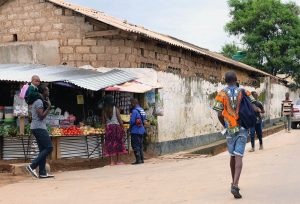 Rauhallinen asvalttipäällysteinen katu Lusakassa Sambiassa, nuori kävelee kadulla reppu selässään, vastaan kävelee 2 nuorta. Kadun laidassa on ruoka- ja vihanneskioski., jonka edessä on 3 aikuista ja yksi pikkulapsi.