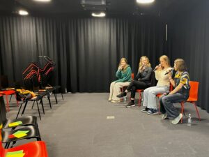 Neljä nuorta henkilöä istuu tummilla verhoilla reunustetussa tilassa ja puhuu mikrofoneihin. Heidän edessään on rivi mustia ja sinisiä tuoleja.