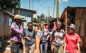 Kuusi nuorta kävelee melkein rivissä kohti kameraa hiekkaisella kujalla, jota reunustavat matalat aaltopellistä rakennetut talot. Nuoret juttelevat ja vitsailevat keskenään.