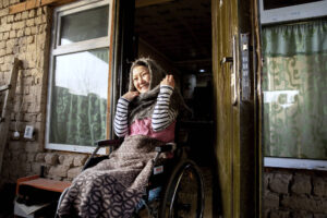 Hymyilevä nainen istuu pyörätuolissa ovensuussa ulkoovella. Hänellä on sylissään viltti ja päänsä ympärille kiedottuna suuri villahuivi. Oven molemmin puolin on ikkunat, jotka heijastavat valoa.