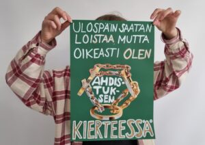 Ruutupaitainen nuori pitää käsissään vihreää julistetta edessään peittäen samalla kasvonsa. Julisteessa on liimattu suuria kultaisten ketjuhen kuvia ja kirjoitettu teksti Ulospäin saatan loistaa, mutta oikeasti olen ahdistuksen kierteessä.