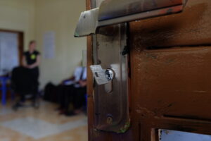 Lähikuvassa raollaan olevan puisen oven metallinen kahva ja lukko. Lukossa on pieni avain. Sisällä huoneessa näkyy epätarkkoina pari ihmishahmoa.