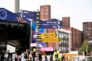 Tolppa keskellä festivaalialuetta. Nuolikylttejä eri suuntiin. Kylteissä lukee mm. info, katuruokaa, Mahdollisuuksien tori.