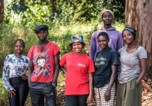 6 nuorta seisoo lähekkäin ulkona aurinkoisena päivänä. Nuoret hymyilevät ja katsovat kuvaajaa kohti. Neljällä on hattu tai pipo päässään. Taustalla näkyy heinikkoa ja puita.