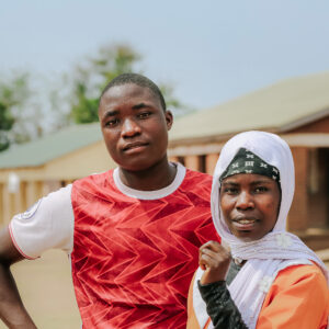 Kaksi nuorta seisoo ulkona lähekkäin ja katsoo kohti. Toisella on punainen t-paita ja toisella päässään valkoinen huivi.