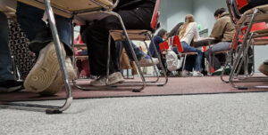 Lattiatasolta kuvattu tuoleja ja pöytiä ryhmissä. Tuoleilla istuu ihmisiä. Kaikilla on jaloissaan tennarit tai lenkkarit.