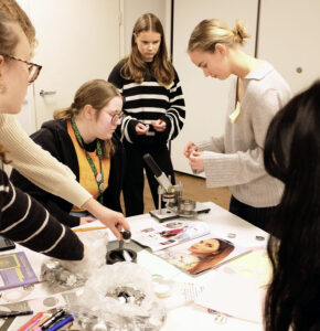 Kuusi henkilöä seisoo tai istuu pöydän ympärillä. Pöydällä on paria, lehtiä, pussillinen metallikiekkoja ja metallinen laite.