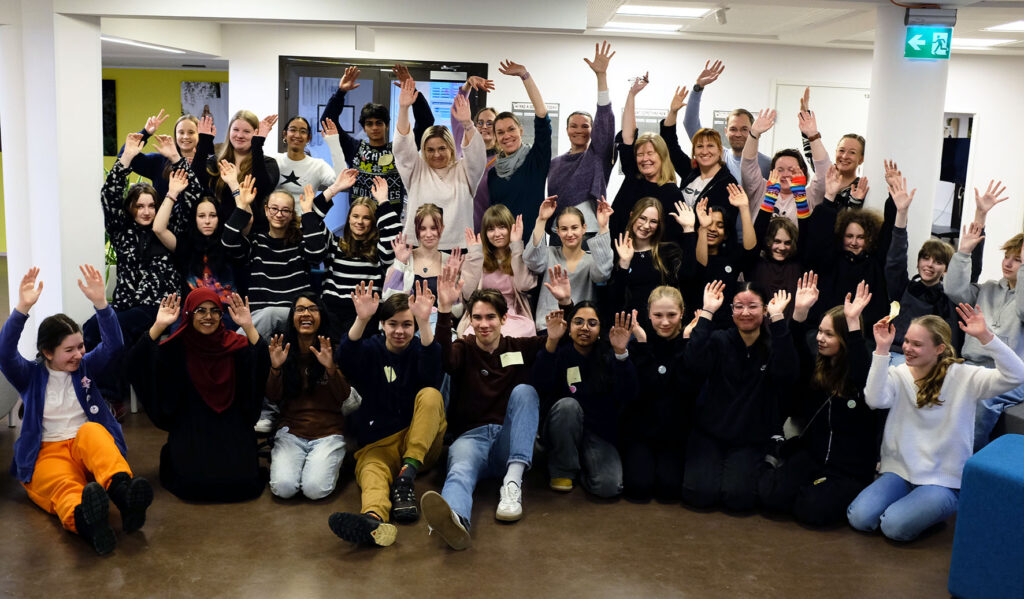 Suuri joukko ihmisiä on nostanut kätensä ilmaan ja nauraa tai hymyilee. Ryhmäkuvassa 27 nuorta ja 9 aikuista.