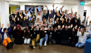 Suuri joukko ihmisiä on nostanut kätensä ilmaan ja nauraa tai hymyilee. Ryhmäkuvassa 27 nuorta ja 9 aikuista.