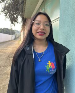 Nuori pitkähiuksinen nainen mustassa takissa ja sinisessä paidassa. Taustalla on vaaleanvihreää seinää ja hiekkainen kuja.