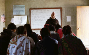 luokkahuoneen edessä opettaja seisoo valkotaulun edessä. 7 nuorta, joista monella on paksu huivi harteillaan, katsoo häntä kohti.