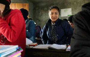 Oppilas pulpetin takana katsoo suoraan kohti. Hänen ympärillään on muita oppilaita, joiden kasvot eivät näy.