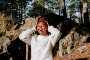 Aggie pitää hiuksistaan kiinni ja hymyilee leveästi kameraan katsoen kallion edessä.