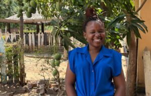 Nuori nainen hymyilee sinisessä paidassa, taustalla on vihreä mangopuu.