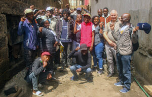 19 ihmistä ryhmäkuvassa kapealla aurinkoisella kujalla kahden kiviseinän välissä.