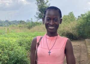 Nuori hymyilevä nainen vaaleanpunaisessa mekossa. Taustalla peltoa.