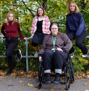 4 nuorta lähekkäin. 1 istuu pyörätuolissa, 2 taaempana metallisella reunakaiteella ja 1 nojaa kaiteeseen.