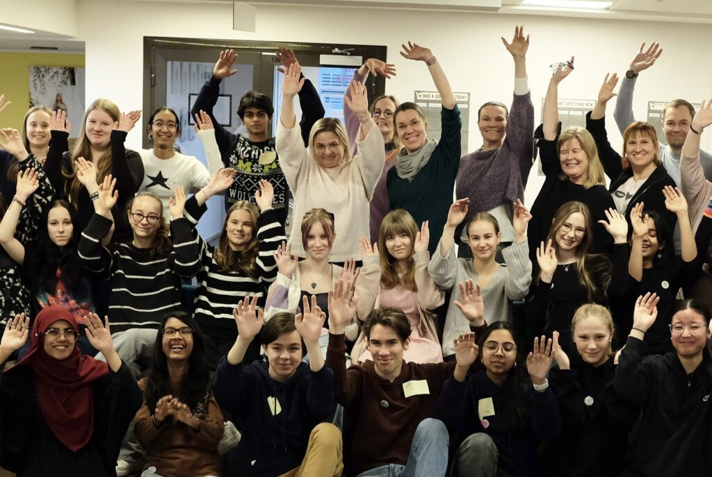 Suuri joukko ihmisiä on nostanut kätensä ilmaan ja nauraa tai hymyilee. Ryhmäkuvassa on 19 nuorta ja 7 aikuista.