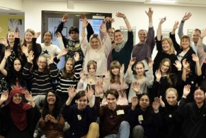 Suuri joukko ihmisiä on nostanut kätensä ilmaan ja nauraa tai hymyilee. Ryhmäkuvassa on 19 nuorta ja 7 aikuista.