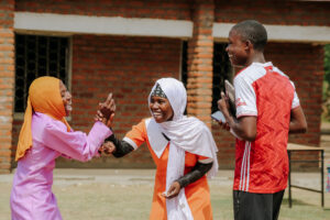 Kolme nauravaa nuorta ulkona. Kaksi lyö oikeat kätensä yhteen. Heillä on kirkkaan väriset pitkähihaiset mekot ja päähuivit. Kolmannella on lyhyet hiukset, punavalkoinen t-paita.