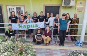16 aikuista ryhmäkuvassa ulkona talon kuistilla, kahdella on valkoinen keppi ja kolme istuu eturivissä maassa. Muutama pitelee banderollia, jossa lukee Kapua.