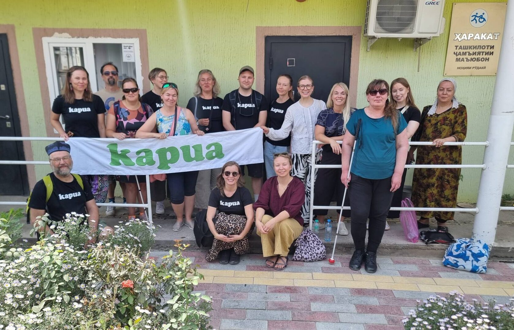 16 aikuista ryhmäkuvassa ulkona talon kuistilla, kahdella on valkoinen keppi ja kolme istuu eturivissä maassa. Muutama pitelee banderollia, jossa lukee Kapua.