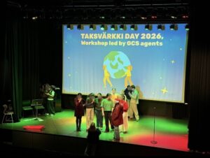 10 nuorta esiintymislavalla, jonka lattia on valaistu erivärisin valoin. Taustalla valkokankaalla piirros maapalloa kantavista ihmishahmoista ja englanninkielinen teksti Taksvärkkipäivä 2026 työpajan ohjaavat Maailmankansalaisen koulun agentit.