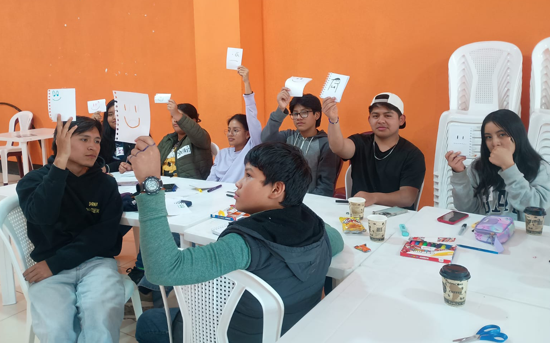 8 nuorta istuu pöytien ääressä. He pitävät ylös nostetussa kädessään valkoisia paperilappuja.