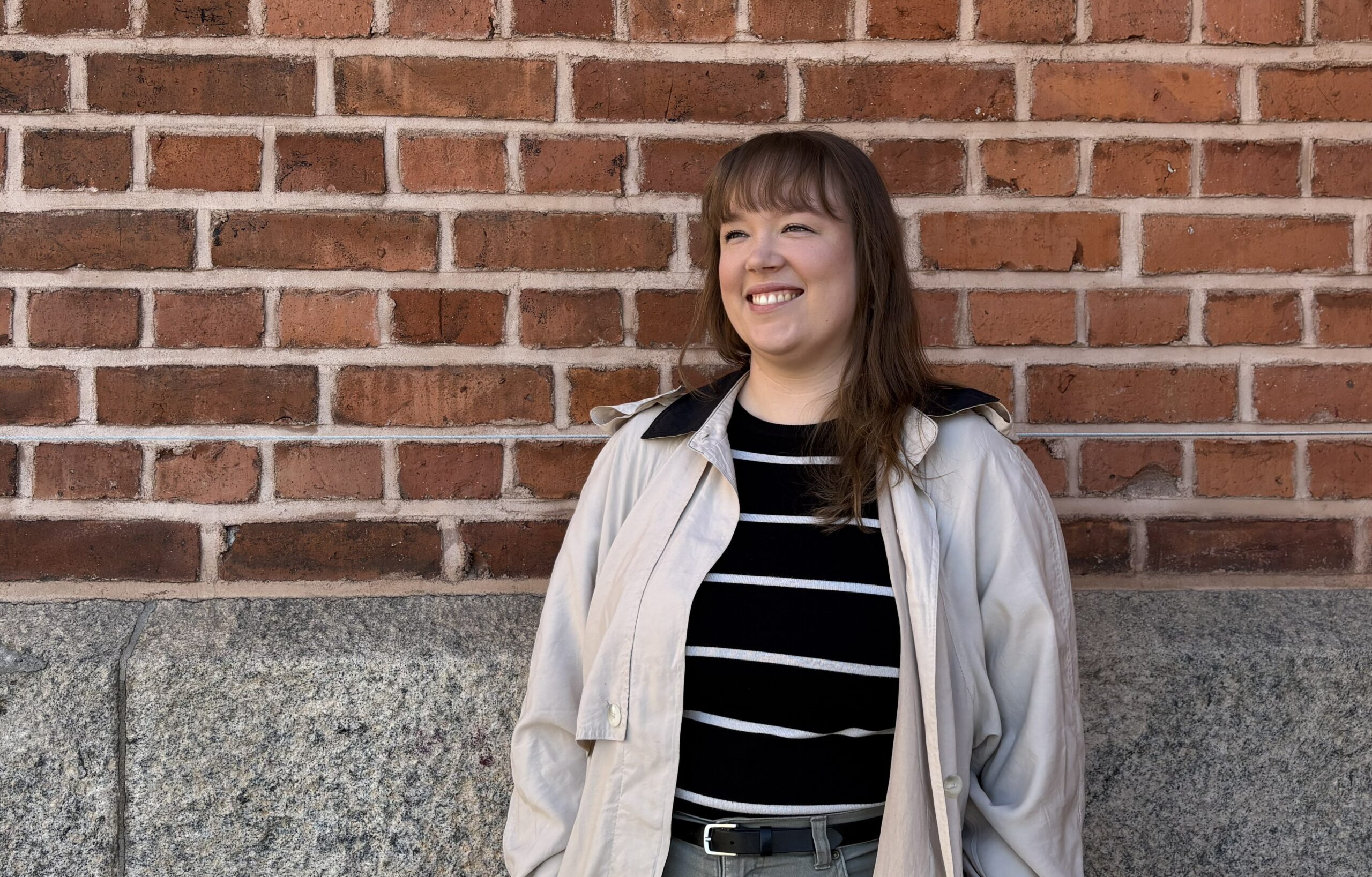 Henkilö seisoo ulkona tiiliseinän edessä. Hän hymyilee ja katsoo viistosti vasemmalle. Henkilöllä on pitkät, ruskeat hiukset, otsatukka ja vaalea takki.