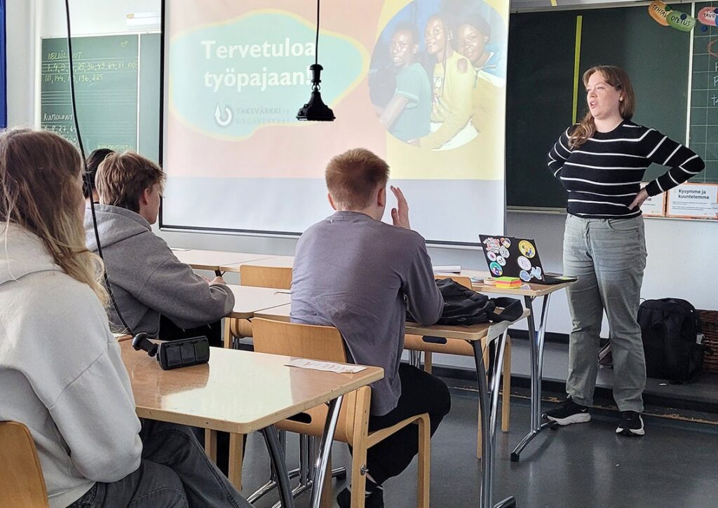 Nuoria luokkahuoneessa kuuntelee edessä seisovaa henkilöä, jonka vieressä valkotaululla lukee Tervetuloa työpajaan.